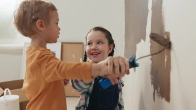 七岁的女孩和两岁的金发男孩拿着滚轴和棕色的粉刷墙。孩子们在微笑。男孩穿着橙色毛衣，女孩穿着方格衬衫