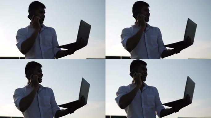 背光镜头，年轻的亚洲工程人员使用笔记本电脑并与智能手机交谈