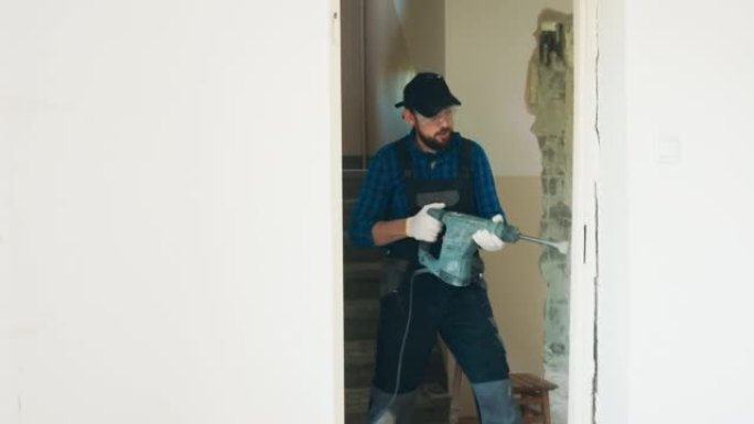 一名男子在使用手持冲击锤翻新公寓时，全力以赴摧毁门框瓷砖。一名戴着安全眼镜的男子穿着工作服大喊