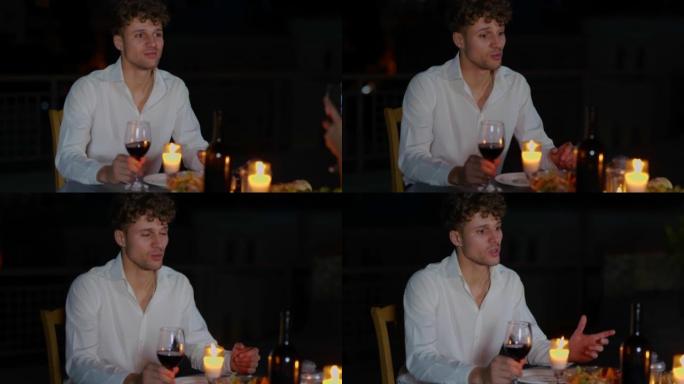 英俊的年轻男性坐在餐桌旁，拿着蜡烛在浪漫的晚餐上，女朋友喝着酒说话。快乐的男人晚上和心爱的女人浪漫约