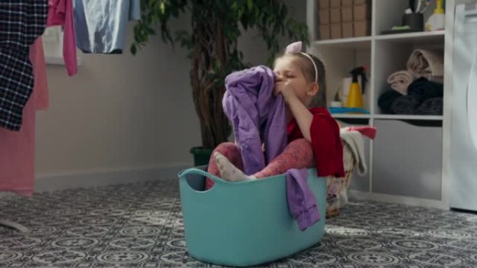 六岁长发女孩坐在蓝色的洗衣碗里。她掏出洗干净的衣服，闻了闻。它闻起来又好又新鲜。洗过的衣服挂在一边。