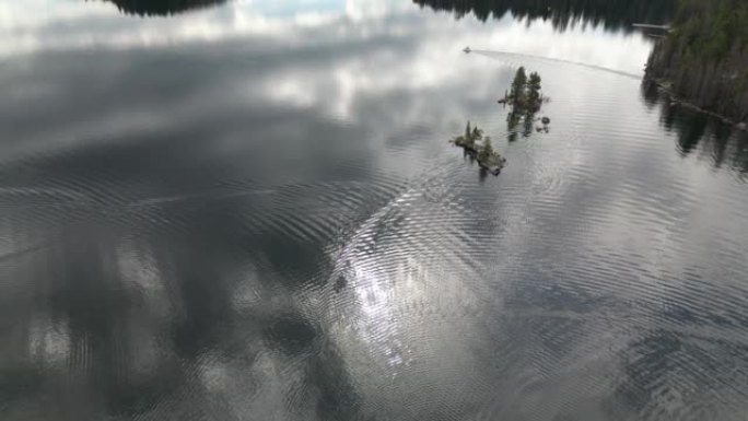 航拍无人机视频，一艘渔船在山湖上行驶，当它驶过水面并在两个小岛周围弯曲时，涟漪从船的后部蔓延开来