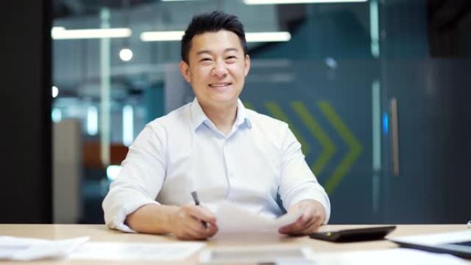 快乐微笑的亚洲员工会计专家的肖像坐在现代办公室的办公桌前，工作场所有文件和笔记本电脑