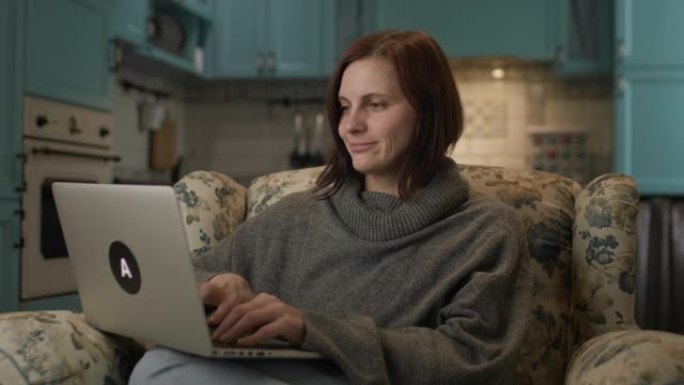 女人在家用笔记本电脑工作。黑发女性坐在扶手椅上在电脑上打字。