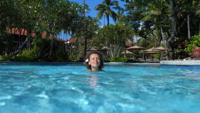梦想旅行目的地。一位美丽的白人妇女喜欢独自在五星级酒店的清水池中游泳。她把脸暴露在阳光下，晒得漂漂亮