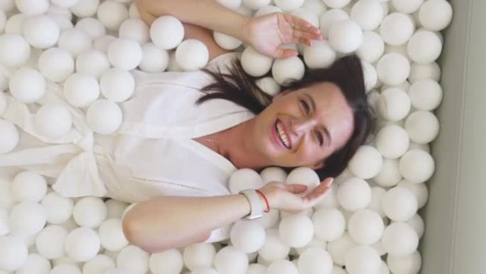 一个穿着白色连衣裙的幸福女人躺在一个装有塑料白球的干燥水池中。