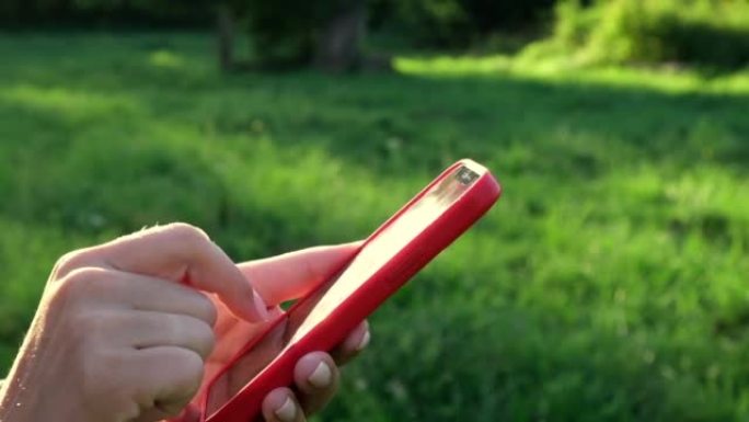 使用智能手机的女性双手特写镜头，自然背景。女人在户外日落时使用手机。无法识别的人触摸智能手机屏幕。不
