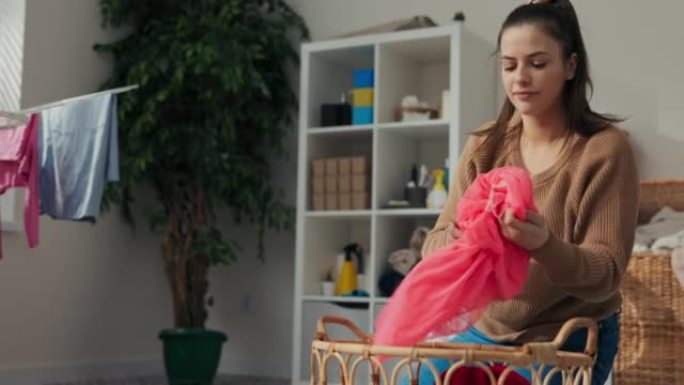 年轻的母亲洗了女儿的衣服，从篮子里拿出来。她折叠袜子和粉红色连衣裙。女人拿着衣服微笑。后面是洗衣机和
