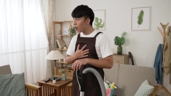 穿着围裙的身体不适的亚洲清洁工男子突然胸痛，正在用吸尘器给客厅的公寓吸尘时，他的心脏试图呼吸