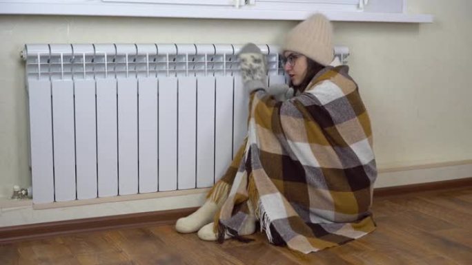 一位年轻女子坐在家里，抱着暖气片，因为天气太冷了。一个戴着帽子和眼镜的女孩坐在地板上，用毯子盖住自己