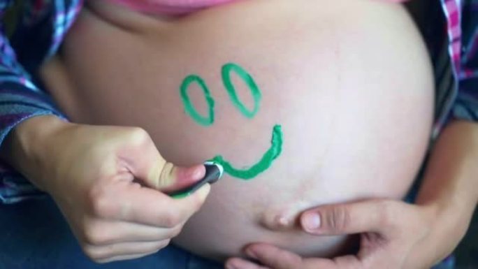 女人在美丽的怀孕肚子上画绿色的微笑，快乐的女性等待她的宝贝孩子的到来，可爱的圆圆的肚子，涂有绿色的眼