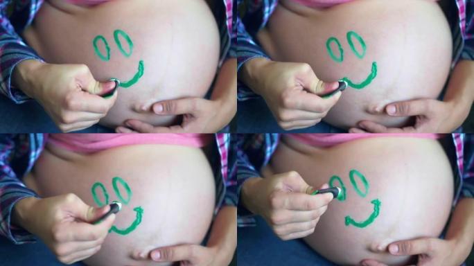 女人在美丽的怀孕肚子上画绿色的微笑，快乐的女性等待她的宝贝孩子的到来，可爱的圆圆的肚子，涂有绿色的眼
