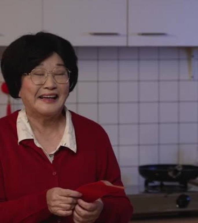 竖屏:中国农历除夕夜，在厨房吃完晚饭后，快乐的亚洲老年母亲尴尬地微笑着接受成年儿媳的压岁钱。翻译:幸