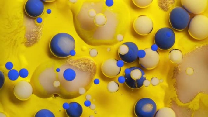 特写彩色金色和蓝色油漆气泡散布在黄色表面上，并混合在抽象设计中。闪闪发光的颗粒，墨球滴和混合。运动中