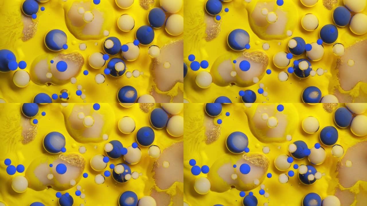 特写彩色金色和蓝色油漆气泡散布在黄色表面上，并混合在抽象设计中。闪闪发光的颗粒，墨球滴和混合。运动中