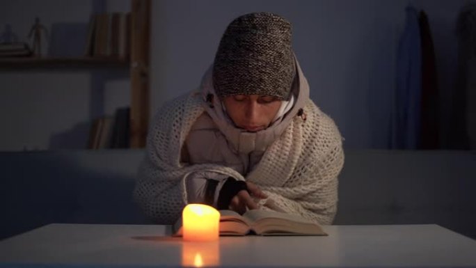 一个穿着温暖的冬帽和围巾的高加索大胡子男人坐在家里的桌子旁，用燃烧的蜡烛温暖双手。家里冬天不供暖的概