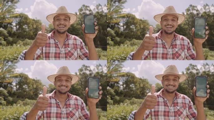 戴着帽子的年轻人农民对相机微笑，并显示带有色度键屏幕的智能手机。拉丁快乐的雄性牧羊人拿着手机演示广告