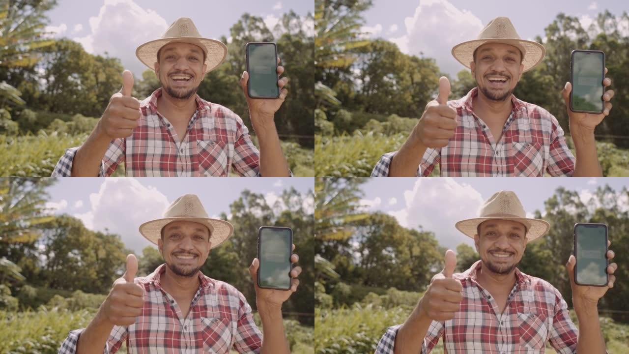 戴着帽子的年轻人农民对相机微笑，并显示带有色度键屏幕的智能手机。拉丁快乐的雄性牧羊人拿着手机演示广告