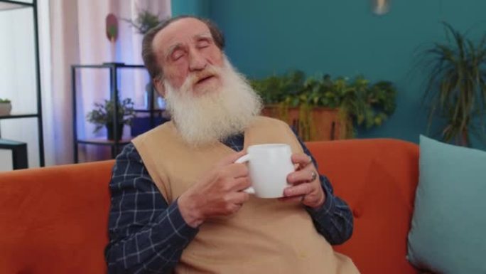 迷人的微笑老爷爷在家里的沙发上喝一杯咖啡或凉茶