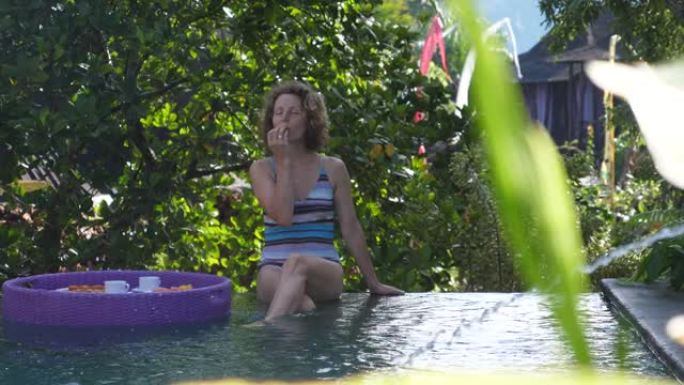 早上好。一位年轻的白人妇女从游泳池里的浮动早餐开始新的一天。她喜欢大自然的声音，喷泉中水的杂音。