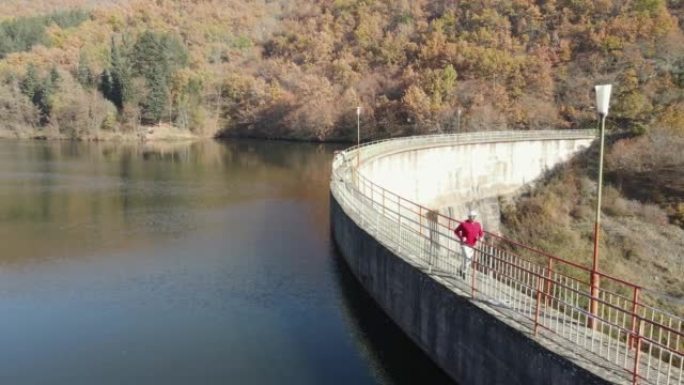 男子在大坝上慢跑，秋天的湖桥，背景是山七彩森林，鸟瞰图，swlo motion
