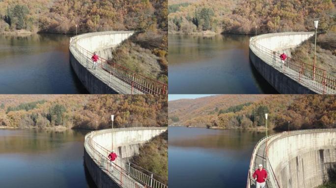男子在大坝上慢跑，秋天的湖桥，背景是山七彩森林，鸟瞰图，swlo motion