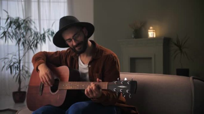 戴着帽子和眼镜的男吉他手e-learning.Man在家里的沙发上弹奏原声吉他。年轻的大胡子家伙在乐