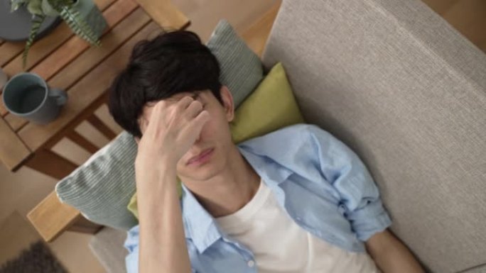 一位疲惫的亚洲年轻男性的俯视图正在家里客厅的沙发上休息时，用手按摩和摩擦他酸痛的眼睛。