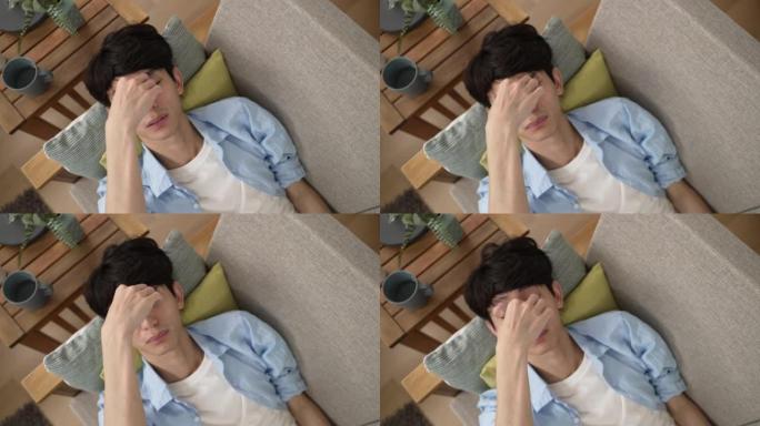 一位疲惫的亚洲年轻男性的俯视图正在家里客厅的沙发上休息时，用手按摩和摩擦他酸痛的眼睛。