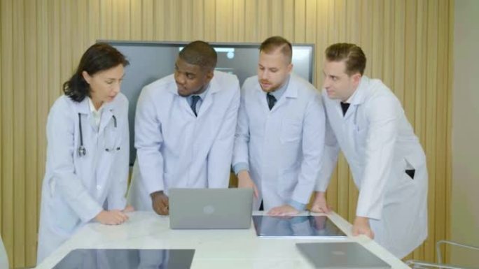 医生团队站在医院会议室，主任医师讨论在笔记本电脑中寻找患者的治疗方法。穿着白大褂的同事一起分析疾病症