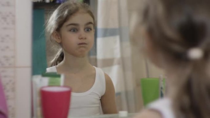 肖像年轻有趣的小女孩喜欢在浴室做鬼脸，看着镜子。快乐肖像可爱的高加索无牙儿童在浴室做鬼脸。有趣的情感