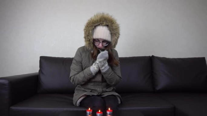 一个穿着夹克的女孩坐在沙发上，桌子上放着蜡烛。一个年轻的女人通过燃烧蜡烛来温暖自己。未付账单。4k