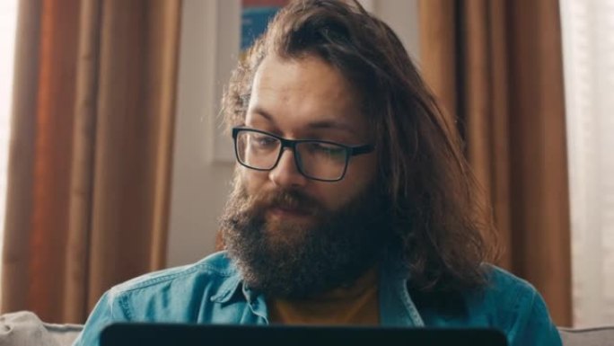 戴着眼镜的大胡子时髦男人的脸的特写镜头在笔记本电脑上浏览互联网，网上商店为亲人寻找礼物，家居饰品