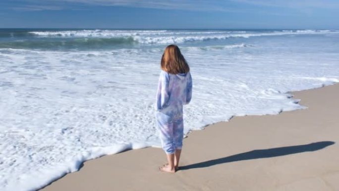 穿着tieday track suite的年轻美女看着大西洋海岸和泡沫的海浪。海滩，蓝天和向远处望去