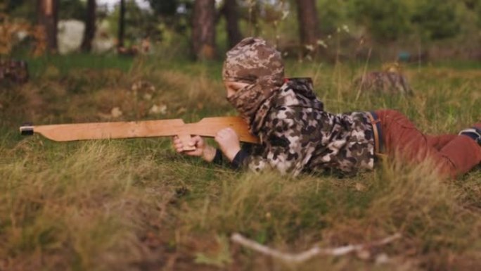身穿迷彩服，头戴巴拉克拉瓦帽，手持木枪的小男孩躺在草地上。年轻的士兵在春天的森林里匍匐前进。侧视图