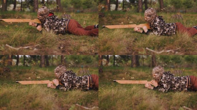 身穿迷彩服，头戴巴拉克拉瓦帽，手持木枪的小男孩躺在草地上。年轻的士兵在春天的森林里匍匐前进。侧视图