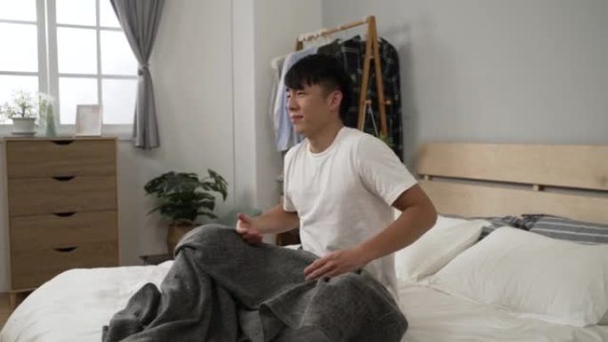昏昏欲睡的亚洲男性坐在床上，早上在明亮的卧室室内起床后，伸懒腰挠头。