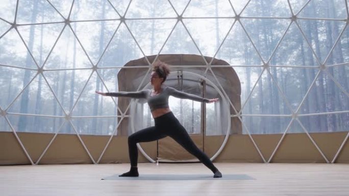 卷发混血女孩练习瑜伽，在概念圆顶工作室做体式。年轻女子穿着黑色灰色运动服站在垫子上练习瑜伽。健身的概