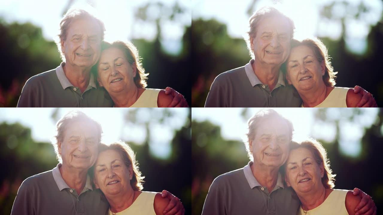 高级夫妇在公园对着镜头微笑。丈夫的肖像脸，胳膊搂着年长的妻子。背光阳光