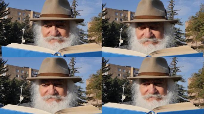 一位老人的肖像，他留着美丽的白胡子，头上戴着帽子，在阳光明媚的日子里在户外的公园里看书。老人手握蓝皮