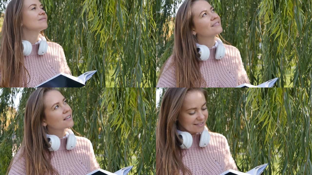 一个可爱的迷人女人站在一棵绿树旁看书，享受着阳光和露天的光芒。在公园看书。美丽的欧洲棕色头发女人