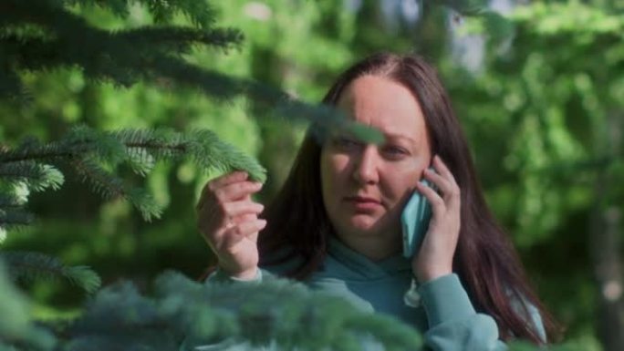 女人正在用手机打电话，站在公园的绿树后面。
