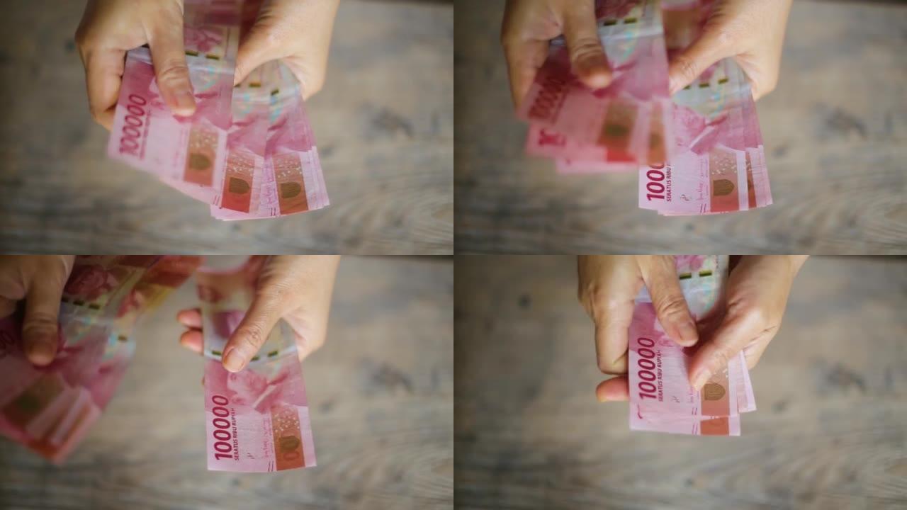 女人数许多印尼盾 (IDR) 红色100,000钞票在手与木制背景。印尼纸币