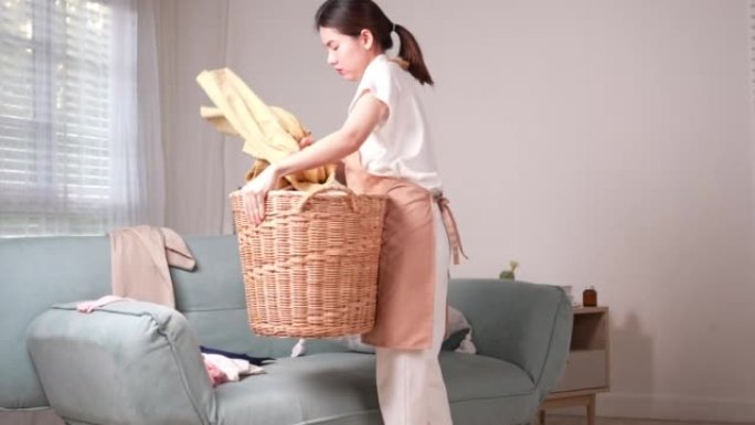 亚洲妇女在家客厅打扫家务。女年轻女管家把凌乱的脏衣服放在沙发上的篮子里做家务。人们呆在家里的概念。