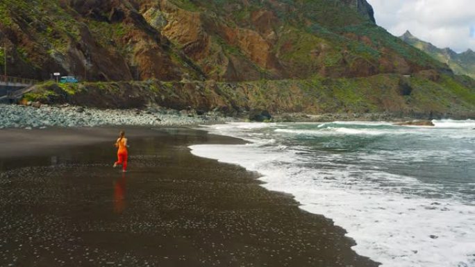 年轻女子在山峦背景下沿着海洋奔跑。夏天的晨光。概念健康户外运动。身体阳性。海浪飞溅泡沫。