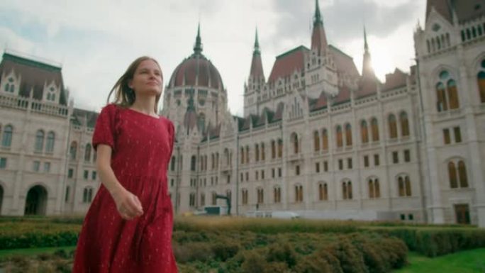在一个阳光明媚的夏日，一名身穿红色连衣裙的女子走在布达佩斯议会大厦附近