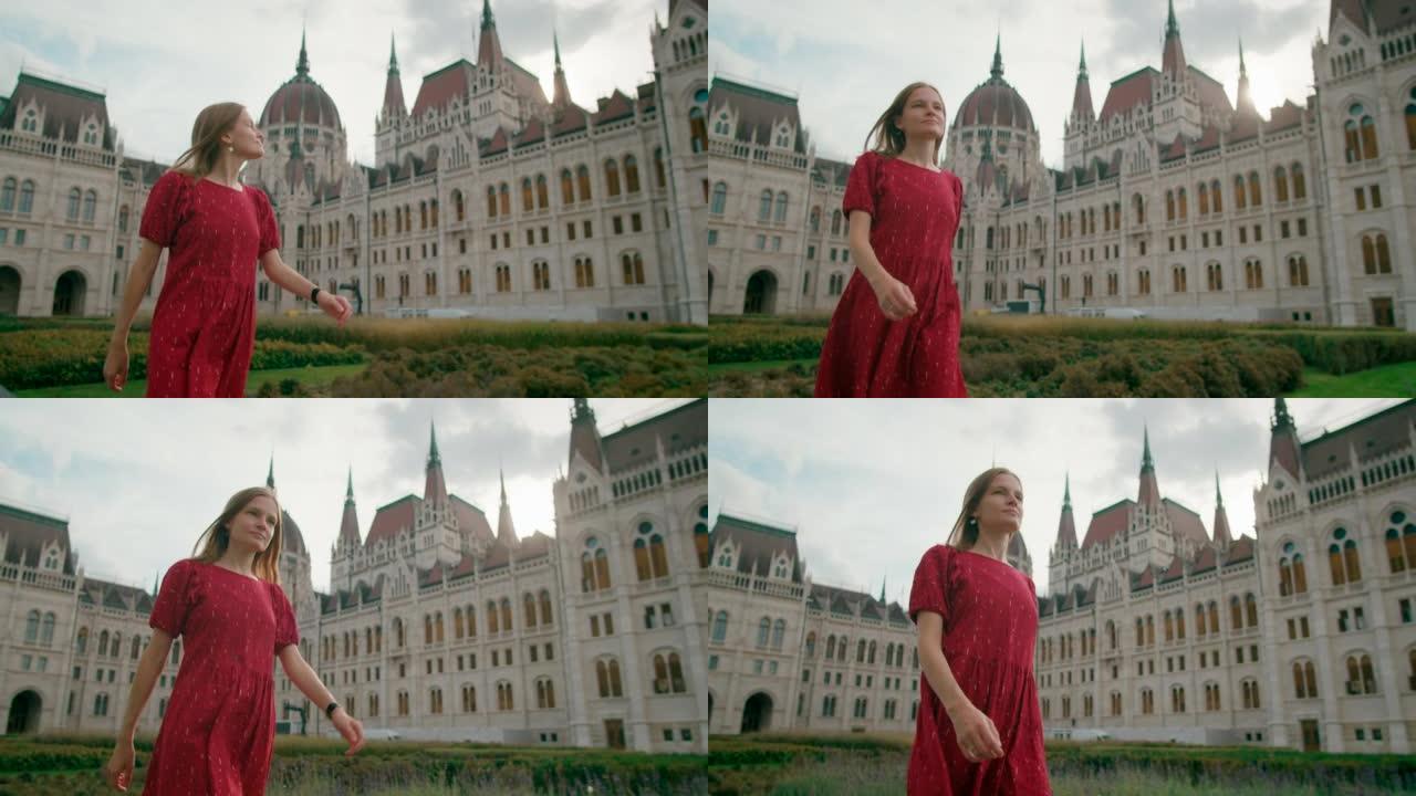 在一个阳光明媚的夏日，一名身穿红色连衣裙的女子走在布达佩斯议会大厦附近