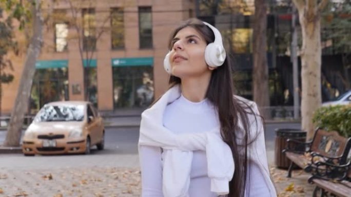 一个年轻的中东长发美丽的女孩在秋天的城市里散步，在她的肩膀上系着一件温暖的白色外套。带着你最喜欢的音