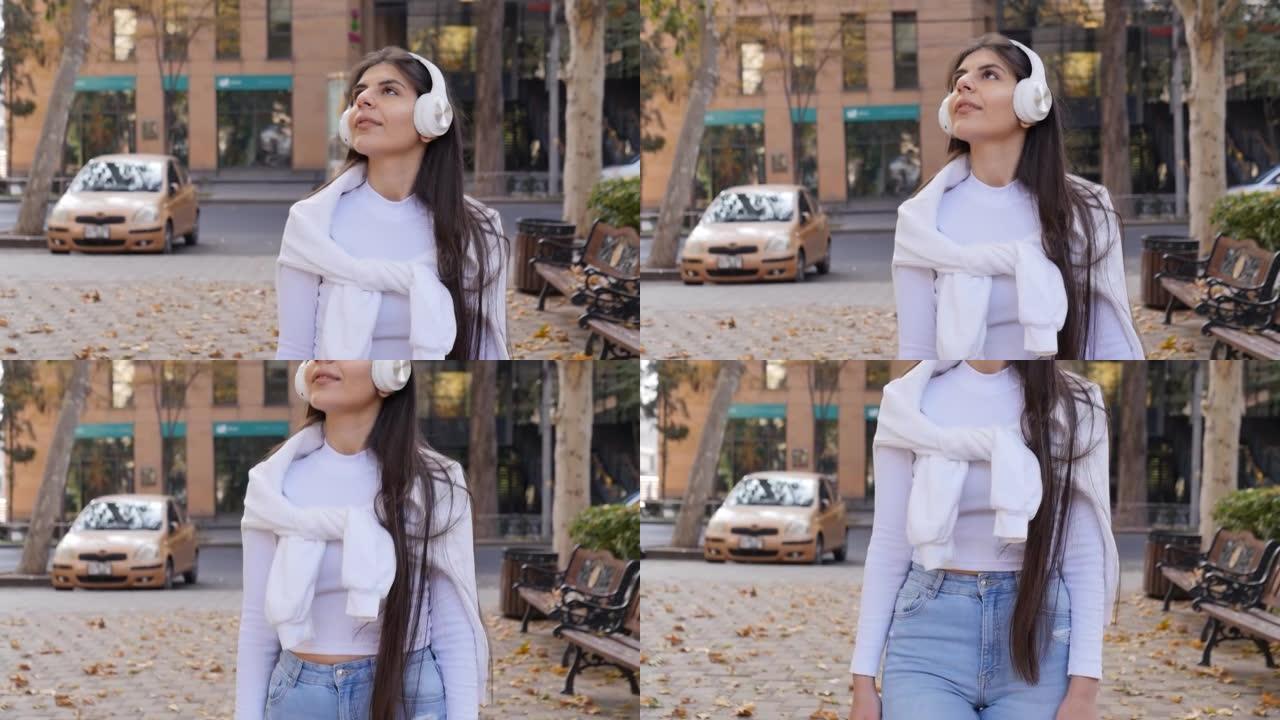 一个年轻的中东长发美丽的女孩在秋天的城市里散步，在她的肩膀上系着一件温暖的白色外套。带着你最喜欢的音