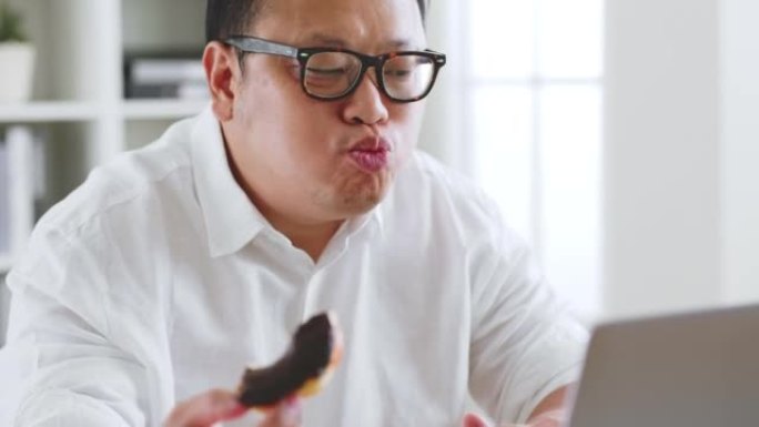 亚洲胖子在使用笔记本电脑时吃甜甜圈，甜食，垃圾食品，不健康的饮食观念
